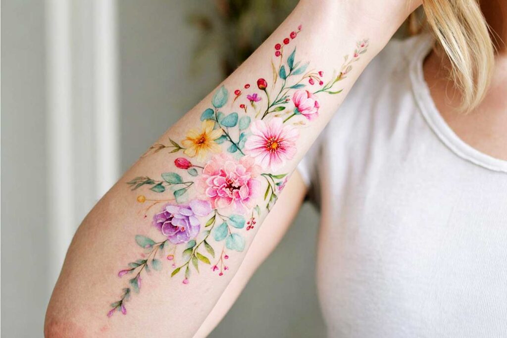Tatuajes pastel, antebrazo interno de una mujer, ramo en cascada de flores silvestres y hojas