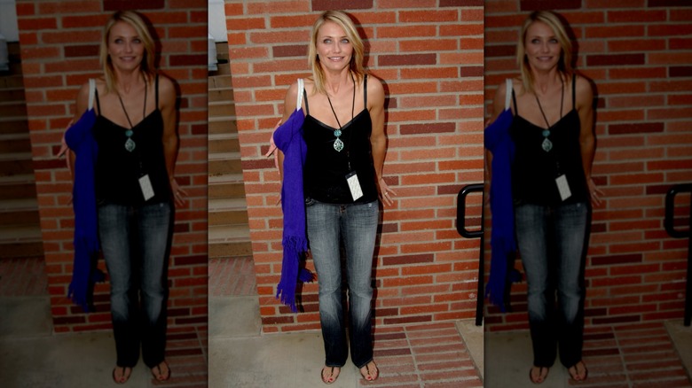 Cameron Diaz con jeans y un top negro de pie junto a una pared de ladrillos