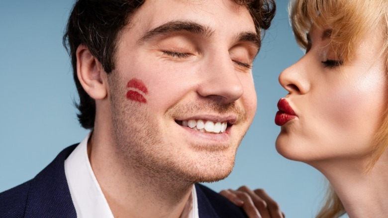 Un hombre con un beso de labios rojos en la mejilla y una mujer fruncida a su lado.