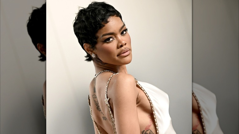 Teyana Taylor posando en un evento de los Oscar 2026. Ella mira por encima del hombro y lleva un vestido de seda color crema sin espalda con detalles en cadenas doradas.