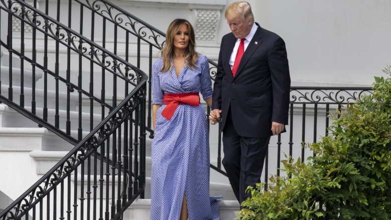 Melania y Donald Trump bajan las escaleras de la Casa Blanca (alejado). Melania lleva un vestido con botones a rayas blancas y azules y un gran cinturón con lazo rojo.