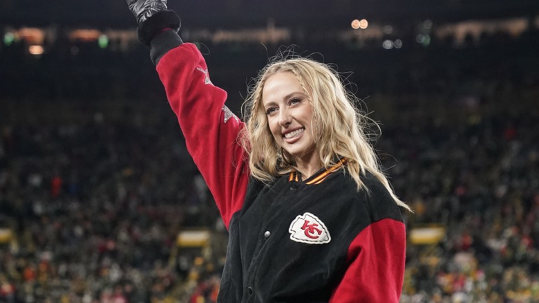 Brittany Mahomes saluda a la multitud en el Super Bowl de 2024. Su cabello rubio está ondulado, con dos pequeñas trenzas en las sienes. Lleva una chaqueta bomber con el tema de los Kansas City Chiefs y un par de guantes de cuero negros.