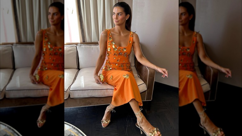 Kaitlan Collins está sentada con las piernas cruzadas en un sofá blanco y tostado. Lleva un vestido naranja cubierto con apliques florales dorados en 3D, tacones de tiras dorados y un mínimo de joyas doradas. Su cabello está peinado hacia atrás detrás de sus hombros.