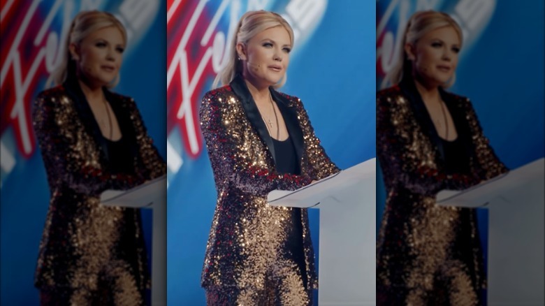 Erika Kirk viste un traje de lentejuelas dorado con solapa negra mientras habla en el podio de un evento de TPUSA. Su cabello rubio está recogido en una coleta alta.