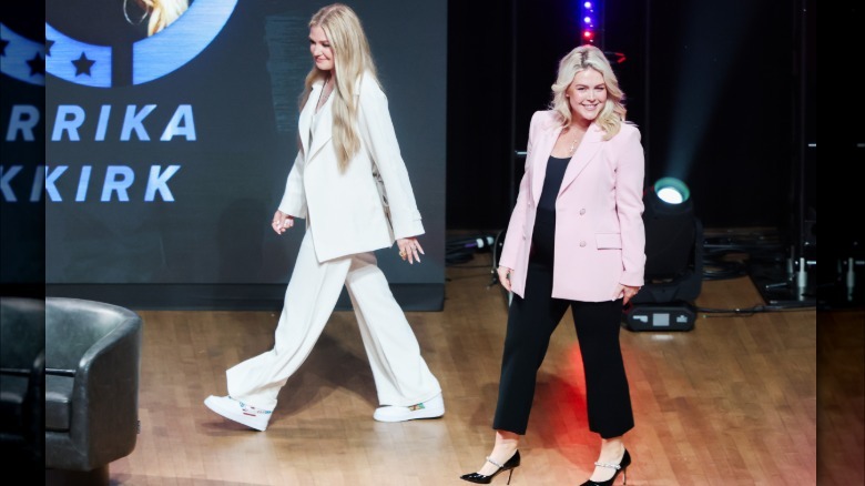 Imagen completa de Kirk y Leavitt en el evento Turning Point USA. Kirk lleva un traje pantalón blanco holgado y zapatillas de deporte. Leavitt lleva una chaqueta rosa claro.
