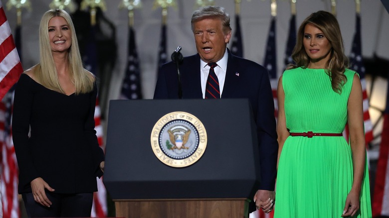 Ivanka y Melania están junto a Donald Trump, un hombre blanco mayor con bronceado en spray y cabello de color claro, en un podio. Ivanka lleva un vestido negro con hombros descubiertos. Melania lleva un vestido verde brillante sin mangas.