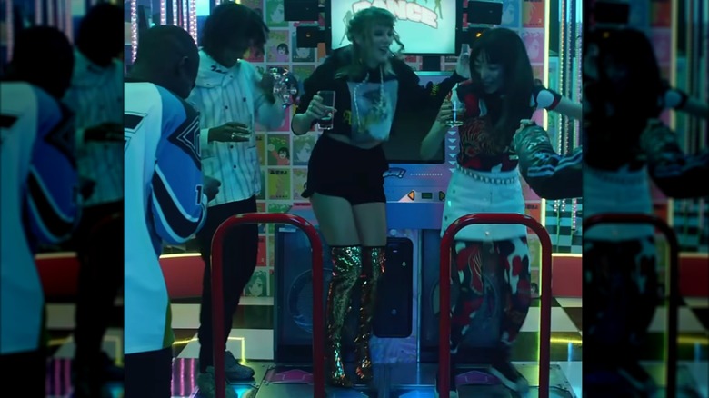 Taylor Swift juega a un juego de baile en una sala de juegos. Lleva una sudadera con capucha corta con gráficos, pantalones cortos negros y un par de botas de lentejuelas doradas por encima de la rodilla.