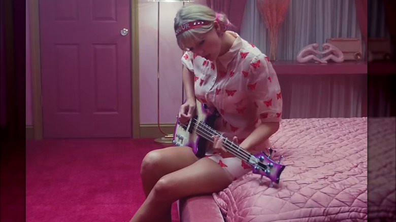 Swift se sienta en un colchón rosa tocando una guitarra rosa. Lleva un vestido rosa claro con botones cubierto de mariposas de color rosa intenso, una diadema de acrílico rosa y cintas rosas para recoger su cabello en dos moños bajos.