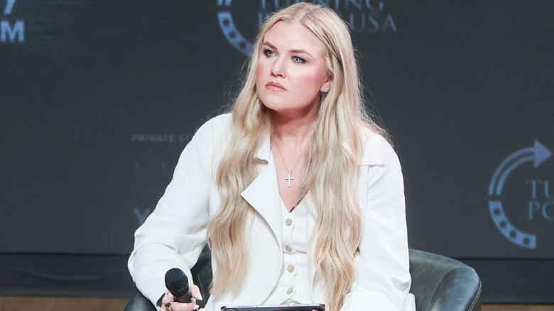 Erika Kirk está sentada en un evento de Turning Point USA. Esta imagen muestra sus extensiones rubias ultralargas. Es una mujer de piel clara y viste una chaqueta de traje blanca y un chaleco a juego.