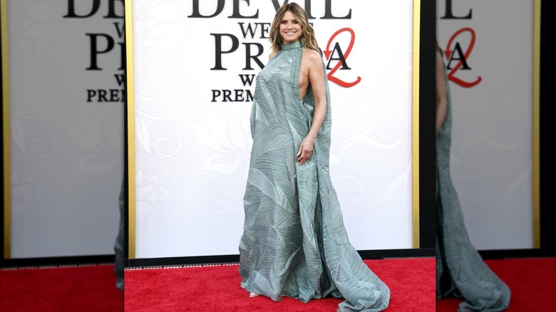 Heidi Klum en el estreno de DWP2. Es una mujer de piel clara y cabello rubio. Lleva un vestido verde espuma de mar, sin mangas, con cuello alto, aberturas laterales reveladoras, diseños de plisados ​​en forma de acordeón y una cola ancha y fluida.