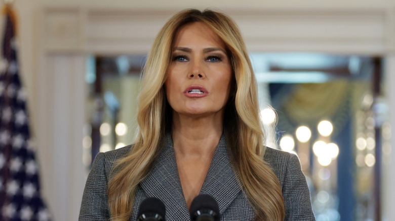 Melania Trump habla en un podio. Ella es una mujer mayor de piel bronceada y cabello rubio. Lleva una chaqueta de traje de tweed negra y gris.