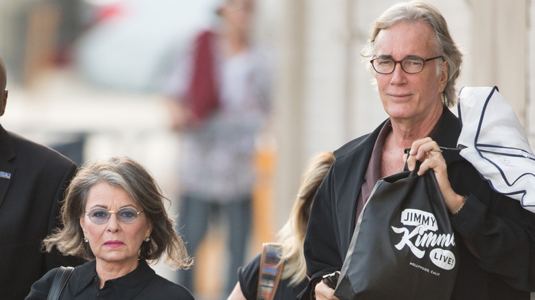 Roseanne Barr camina junto a su compañero Johnny Argent con una expresión severa en su rostro.