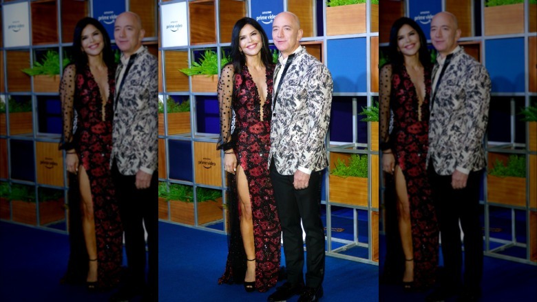 Imagen completa de Lauren Sánchez Bezos y Jeff Bezos. Lauren lleva un vestido largo de cuentas rojas y negras con escote alto y escote bajo. Jeff lleva una chaqueta de traje estampada y pantalones negros.