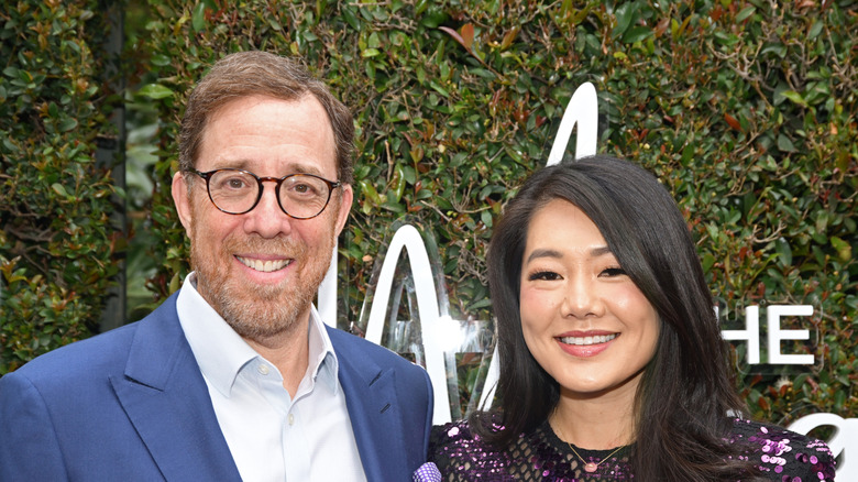 Rob Minkoff y Crystal Kung sonríen mientras posan juntos frente a un fondo de follaje verde