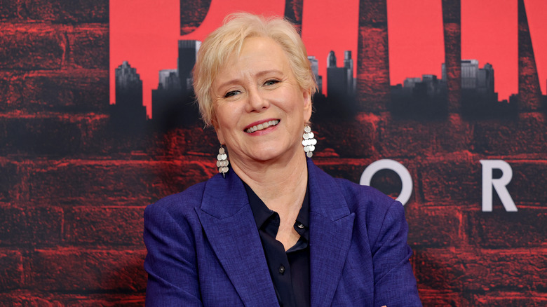 Eve Plumb usa una chaqueta azul y aretes colgantes mientras posa frente a un fondo rojo.
