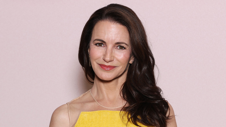 Kristin Davis usa un vestido amarillo brillante y un labio rojo llamativo mientras posa frente a un fondo rosa claro.