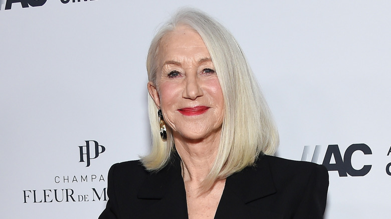 La actriz Helen Mirren sonríe mientras usa chaqueta negra y lápiz labial rojo en un evento.