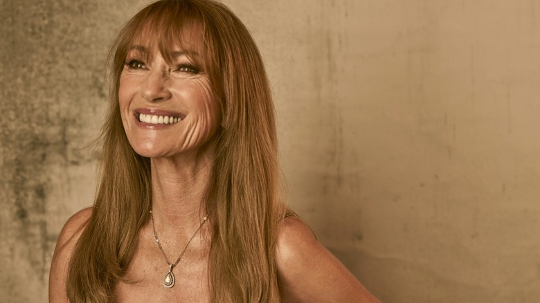 Jane Seymour sonriendo y luciendo un collar con colgante sobre un fondo marrón.