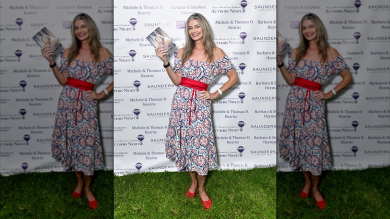 Paulina Porizkova parada sobre el césped sosteniendo un libro y usando zapatos rojos y un vestido en un evento.