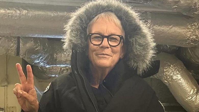 Jamie Lee Curtis haciendo un signo de la paz mientras usa un abrigo con capucha y ribete de piel.