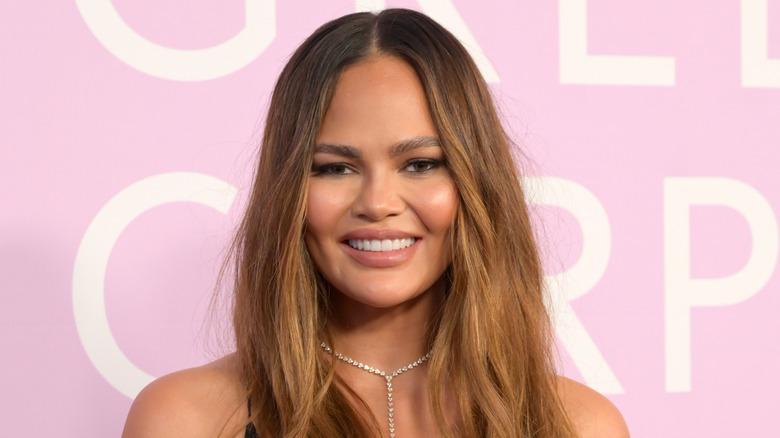 Chrissy Teigen sonríe en un evento de alfombra roja luciendo un collar de diamantes sobre un fondo rosa.