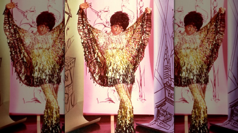 Diana Ross posando dramáticamente con los brazos en alto luciendo un traje dorado brillante.