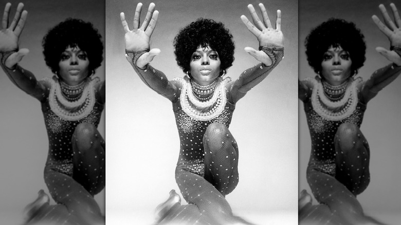 Diana Ross posando para un retrato en blanco y negro con las manos hacia adelante y luciendo un mono de rejilla negro cubierto de pedrería.