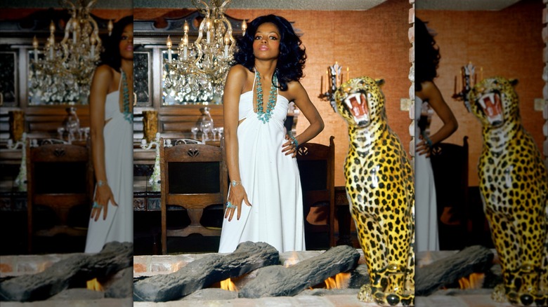 Diana Ross posando junto a una estatua de guepardo con un vestido blanco fluido y un llamativo collar verde.