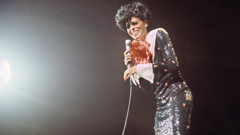 Diana Ross fotografiada en el escenario con un micrófono y luciendo un vestido de lentejuelas negro con un amplio cuello blanco.