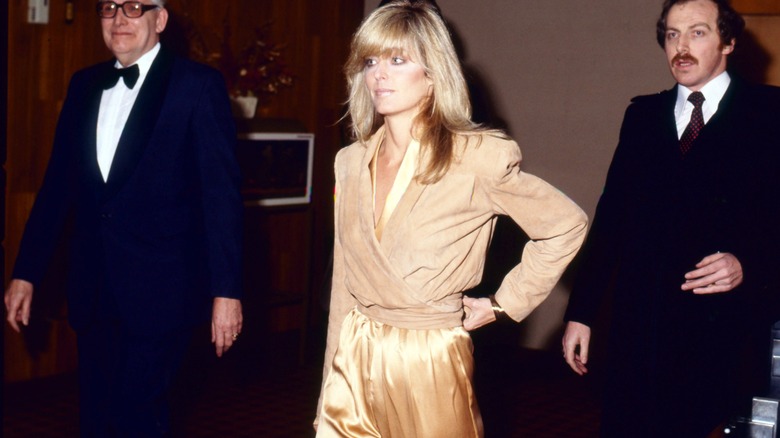 Farrah Fawcett lleva una chaqueta beige y un mono dorado.
