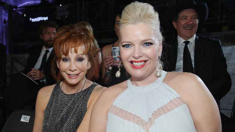 Reba McEntire con un vestido negro brillante y Melissa Peterman con un vestido blanco sentadas juntas.