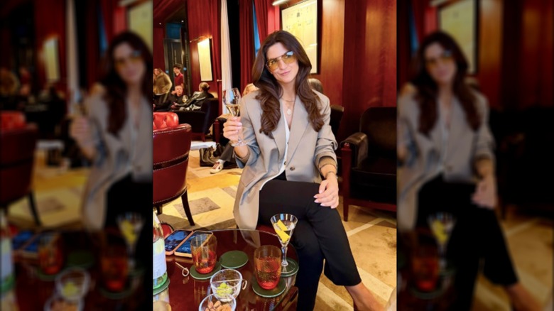 Kaitlan Collins vestida con chaqueta color canela y pantalones con gafas de sol amarillas sosteniendo una copa de champán.