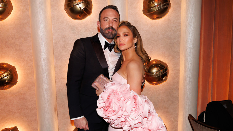 Ben Affleck con traje y Jennifer López con un vestido rosa con volantes posando juntos en un evento de alfombra roja.