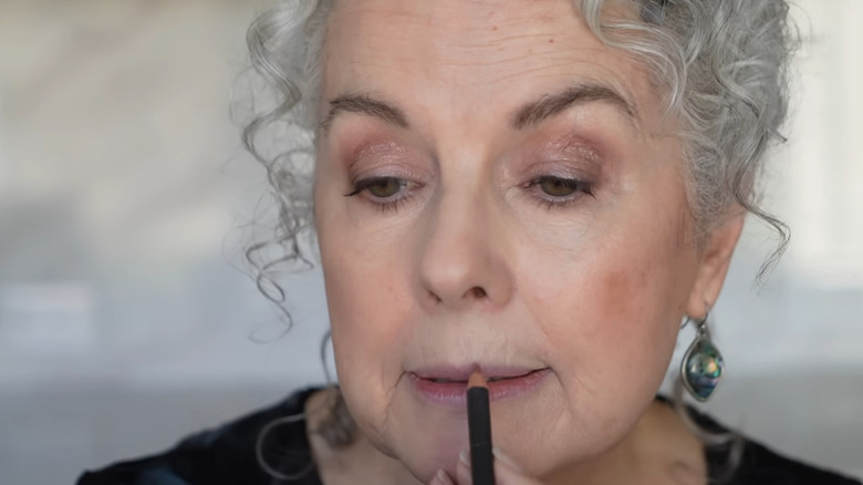 Mujer mayor con cabello gris rizado aplicando delineador de labios