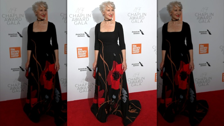 Helen Mirren posando en la alfombra roja en 2018 luciendo un vestido largo negro con flores rojas.