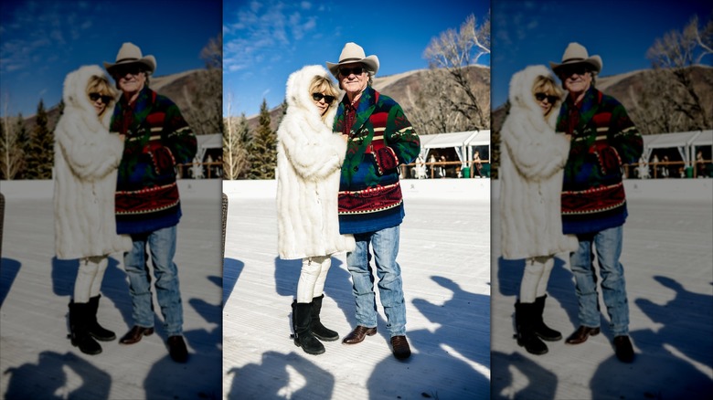Goldie Hawn con un abrigo de piel blanco se toma del brazo con Kurt Russell con un suéter multicolor y jeans.