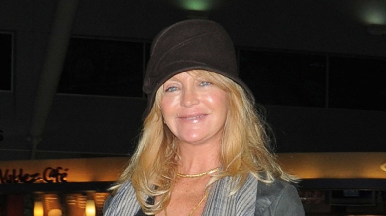 Goldie Hawn sonriendo con un sombrero negro y una chaqueta gris con collares dorados.