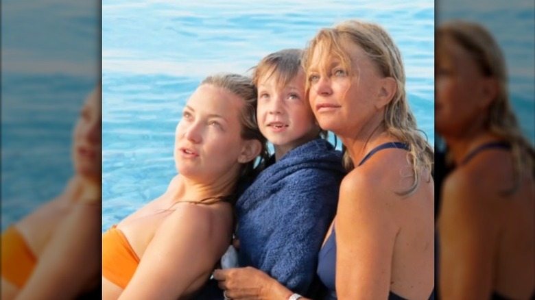 Kate Hudson, su hijo Ryder y Goldie Hawn posan mirando hacia arriba junto al agua en traje de baño.