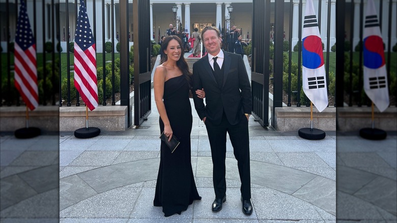 Chip Gaines con traje y Joanna Gaines con un vestido negro sin tirantes posando frente a banderas.