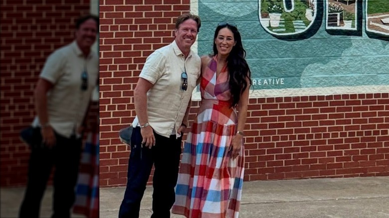 Chip Gaines con una camisa blanca abotonada y Joanna Gaines con un vestido a cuadros posando juntos frente a una pared de ladrillos rojos.