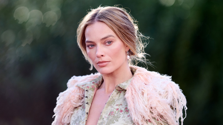Margot Robbie posando afuera en un evento con el cabello recogido y luciendo un conjunto detallado y difuso.