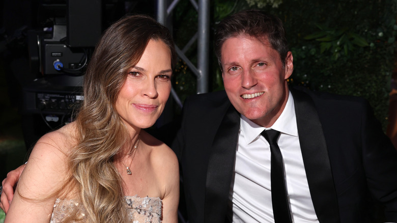 Hilary Swank sonriendo con un vestido sin tirantes junto a su marido Philip Schneider con traje.