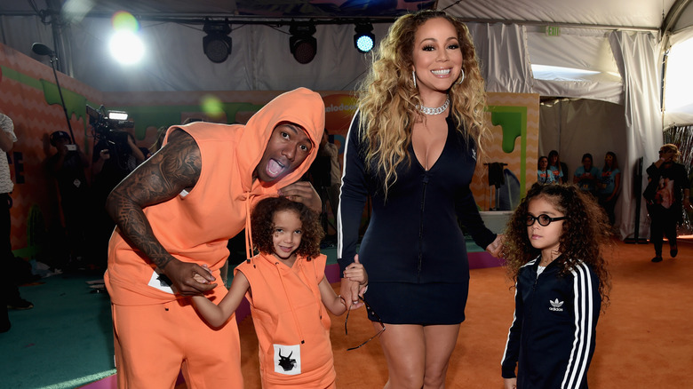 Nick Cannon con un conjunto con capucha naranja y Mariah Carey con un vestido corto negro sonriendo en la alfombra roja con los niños Marruecos y Monroe.