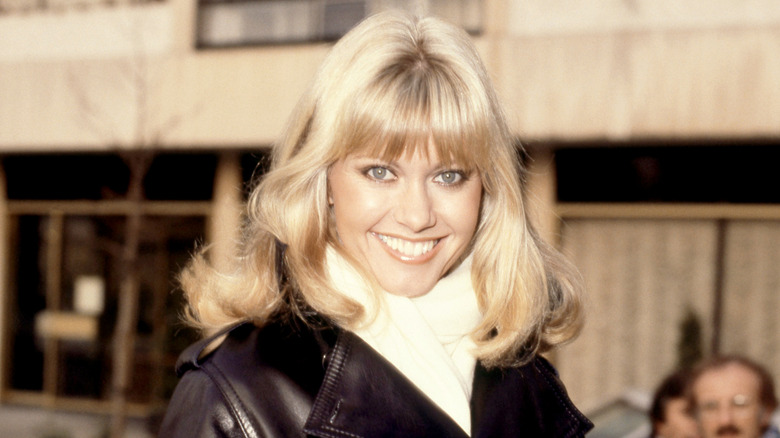 Olivia Newton-John sonríe para los fotógrafos con una chaqueta de cuero negra y una bufanda de lana blanca