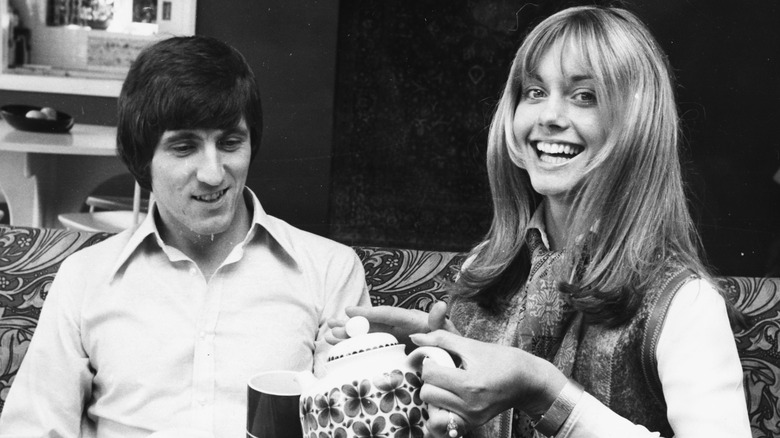 Se ve a Bruce Welch y Olivia Newton-John tomando té juntos en un sofá en una foto en blanco y negro.