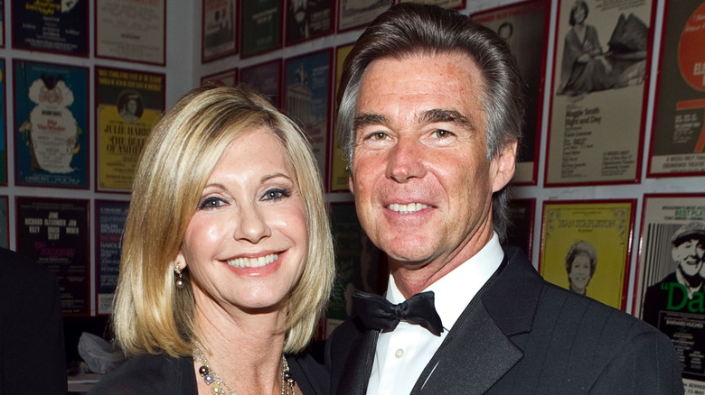Olivia Newton-John sonríe mientras posa junto a su segundo marido, John Easterling, frente a la pared de una galería de carteles.