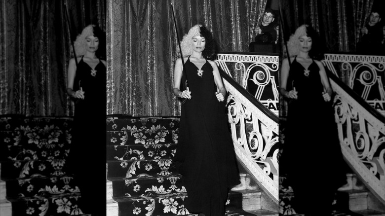 Bianca Jagger bajando unas escaleras ornamentadas con un vestido negro con un bastón y el pelo partido en blanco y negro.