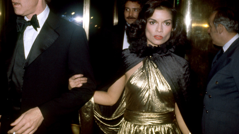 Bianca Jagger luciendo un vestido dorado y una capa negra en un evento nocturno.