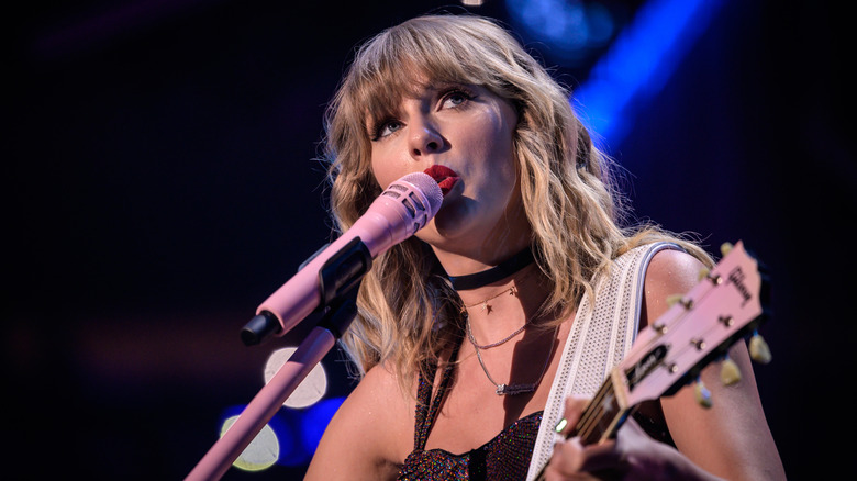 Taylor Swift usa lápiz labial rojo brillante mientras canta frente a un micrófono rosa y toca la guitarra en el escenario.