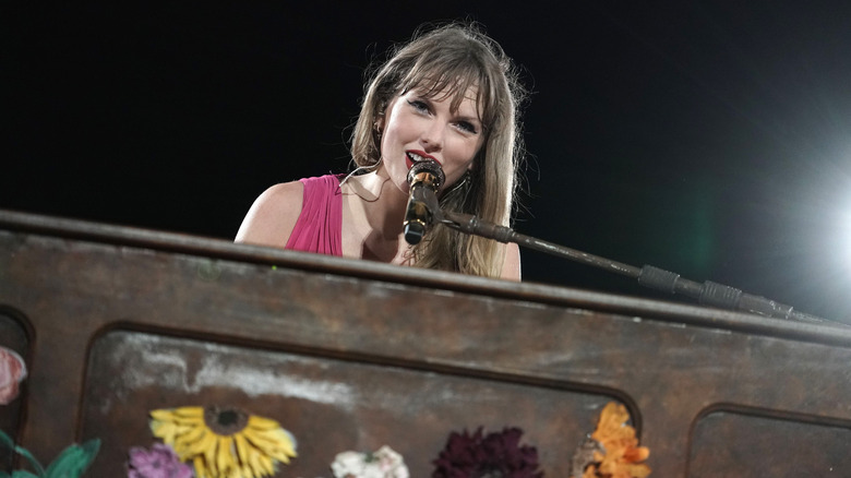 Taylor Swift sonríe hacia la cámara mientras está sentada frente al piano y cantando frente a un micrófono.